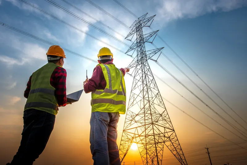 Deux ingénieurs en équipement de sécurité inspectant des lignes électriques à haute tension au coucher du soleil.